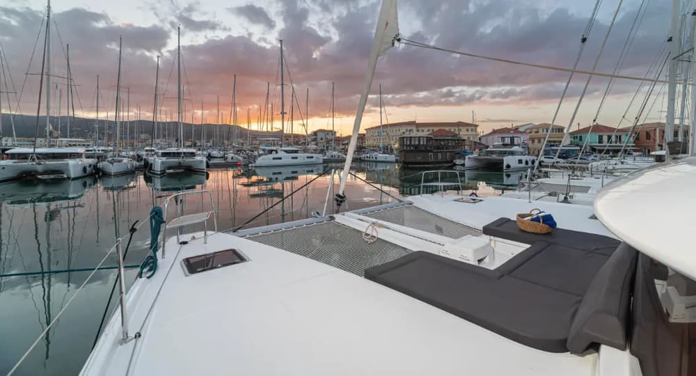 Lagoon 46 catamaran front deck lounge area with cushioned sunbeds, overlooking a scenic marina at sunset – a perfect spot for relaxation on a sailing holiday.