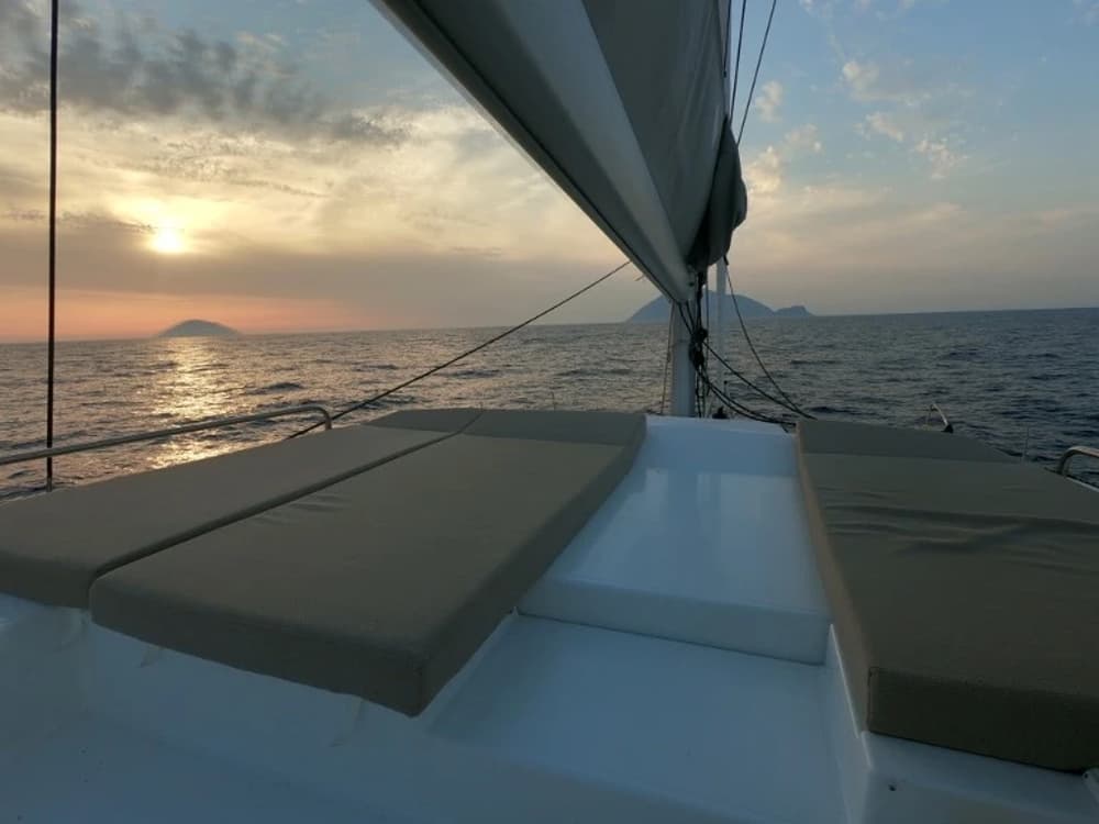 Spacious forward deck lounge of a catamaran with cushioned sun pads, offering a comfortable relaxation area while sailing at sunset.