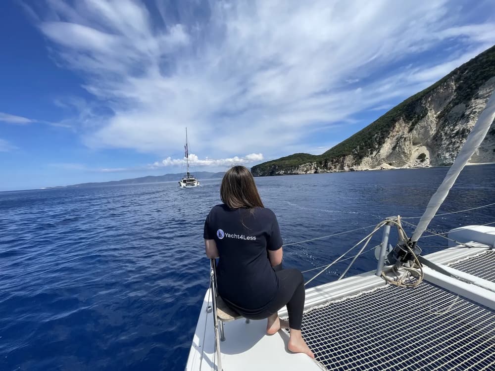 Yacht4Less guest relaxing on a catamaran trampoline with panoramic views of the Ionian coastline and sailing yachts ahead