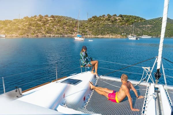 A couple relaxing on the netted bow of a catamaran anchored in a tranquil bay, surrounded by scenic greenery and calm waters, representing the freedom of a bareboat charter.