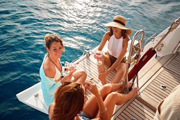 Three women enjoying glasses of wine on the deck of a yacht, surrounded by calm blue waters, illustrating the social and relaxing atmosphere of a cabin charter holiday.