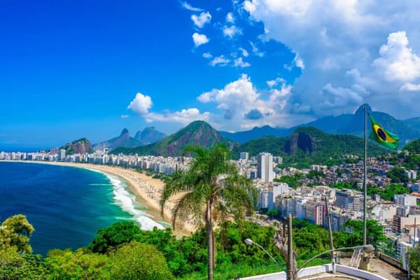A panoramic view of Rio de Janeiro's Copacabana Beach with the Brazilian flag, lush greenery, and iconic mountains in the background, showcasing the vibrant beauty of sailing holidays in Brazil.