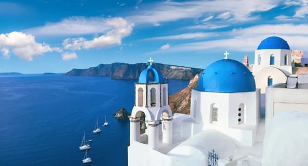 The iconic blue-domed churches of Oia, Santorini, overlooking the Aegean Sea with anchored sailboats below, showcasing the beauty of sailing holidays in Greece.