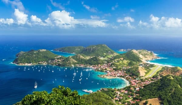 An aerial view of the beautiful coastline of Guadeloupe, featuring sailboats anchored in the clear turquoise waters, with lush hills and vibrant villages, highlighting the scenic allure of sailing holidays in the Caribbean.