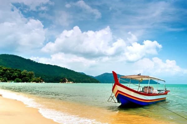 A traditional wooden boat anchored on a sandy beach with turquoise waters and lush green hills in the background, showcasing the scenic beauty of sailing holidays in Malaysia