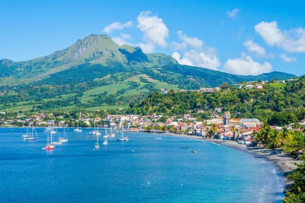 A picturesque view of a coastal town in Martinique with anchored sailboats, lush green mountains, and a vibrant shoreline, showcasing the charm of sailing holidays in the Caribbean.