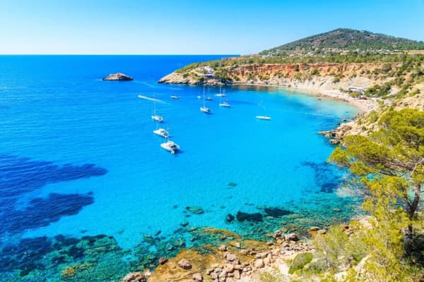 A picturesque bay in Ibiza, Spain, with turquoise waters, anchored sailboats, and rugged cliffs, showcasing the vibrant beauty of sailing holidays in the Balearic Islands.