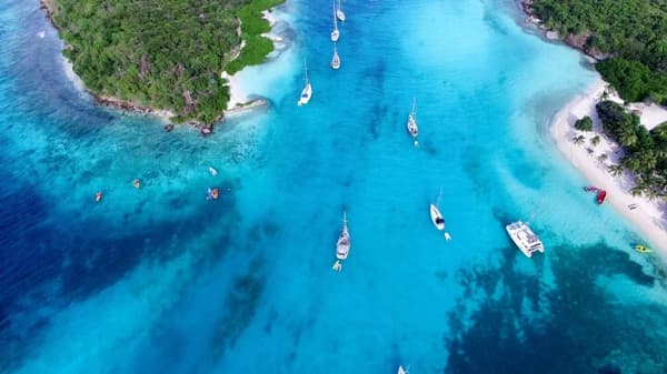 An aerial view of sailboats anchored in crystal-clear turquoise waters near a lush green island in Saint Vincent, showcasing the serene beauty of sailing holidays in the Caribbean.