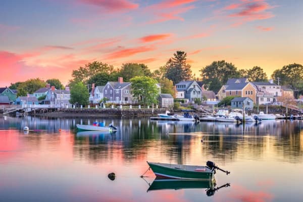 A picturesque waterfront village in New England at sunset, with small boats reflected in calm waters, capturing the charm and serenity of sailing holidays in the USA.