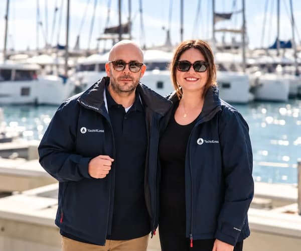 Yacht4Less charter team members wearing branded sailing jackets at a marina with yachts